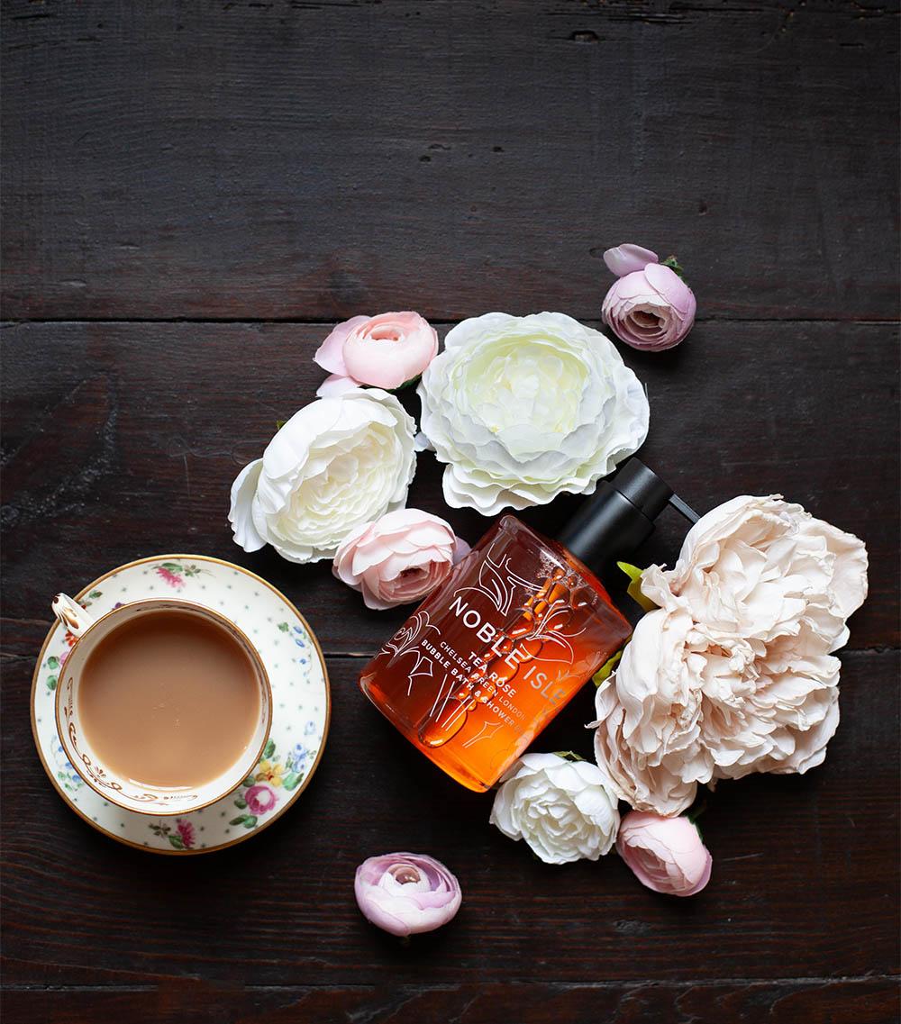 Flowers and a noble isle tea rose bath & shower gel bottle lay down on a black wooden background with a cup of tea in bone china